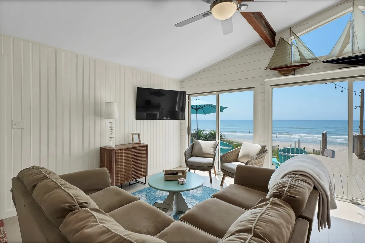 This image depicts a bright and inviting living area, featuring a comfortable sofa, a coffee table, and large windows showcasing a stunning view of the beach. Nautical decorative accents, including sailboats, reinforce the coastal setting. The space feels open and tranquil, perfect for unwinding after a day at the shore.