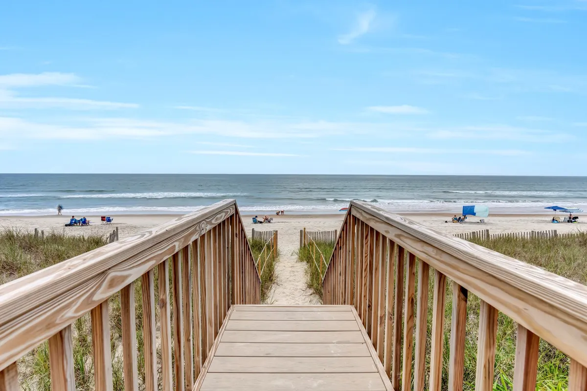 A wooden walkway leads from the cottage over the dunes directly onto the sandy beach, with distant figures enjoying the water and scattered beach chairs. The scene captures the seamless transition from cottage to shore.