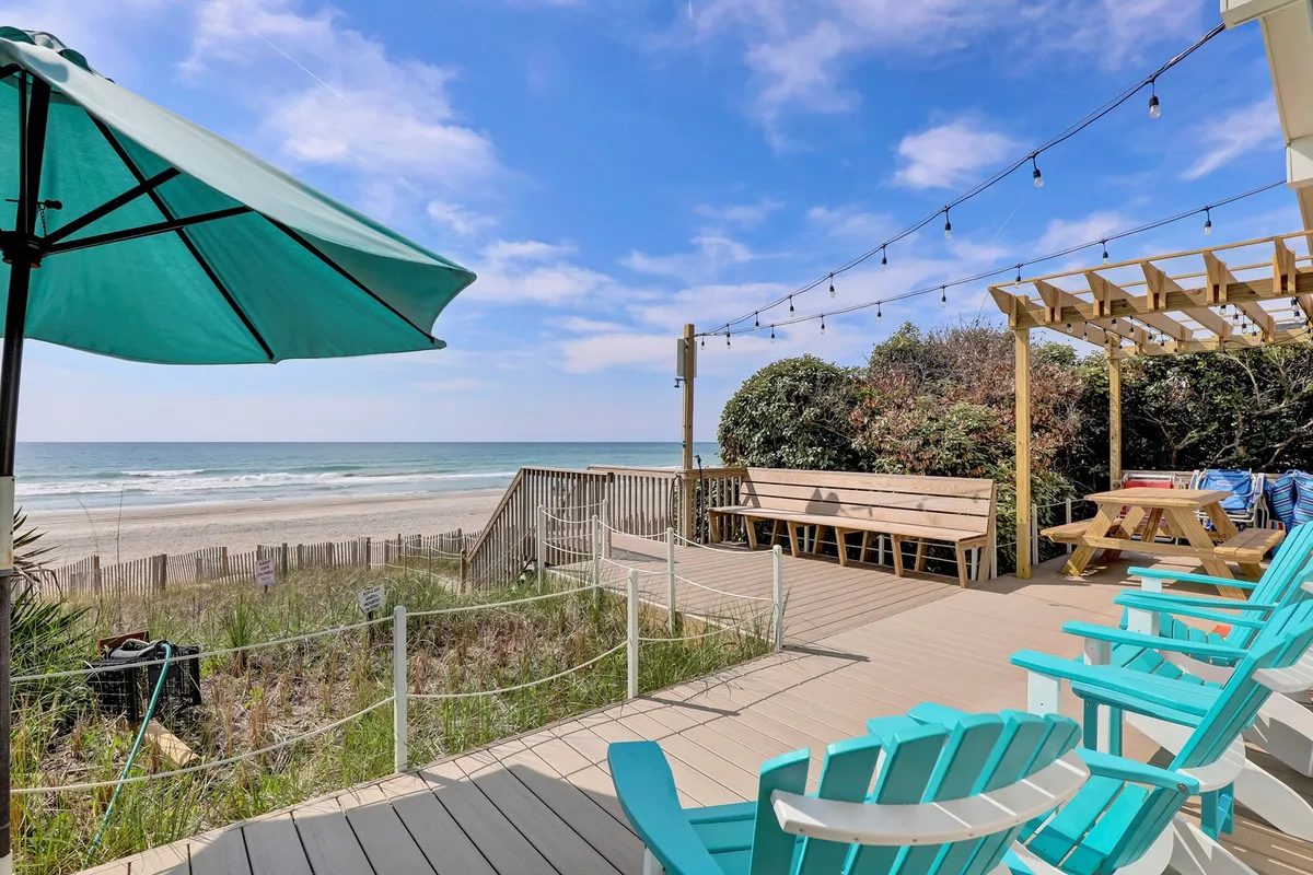 The image shows a cozy beachfront cottage with a spacious deck overlooking the ocean. Deck chairs and a welcoming outdoor seating area are arranged for relaxation and socializing, while string lights hint at magical evenings under the stars. The setting is bright and cheerful, capturing the laid-back vibe of Surf City and offering panoramic sunset views.