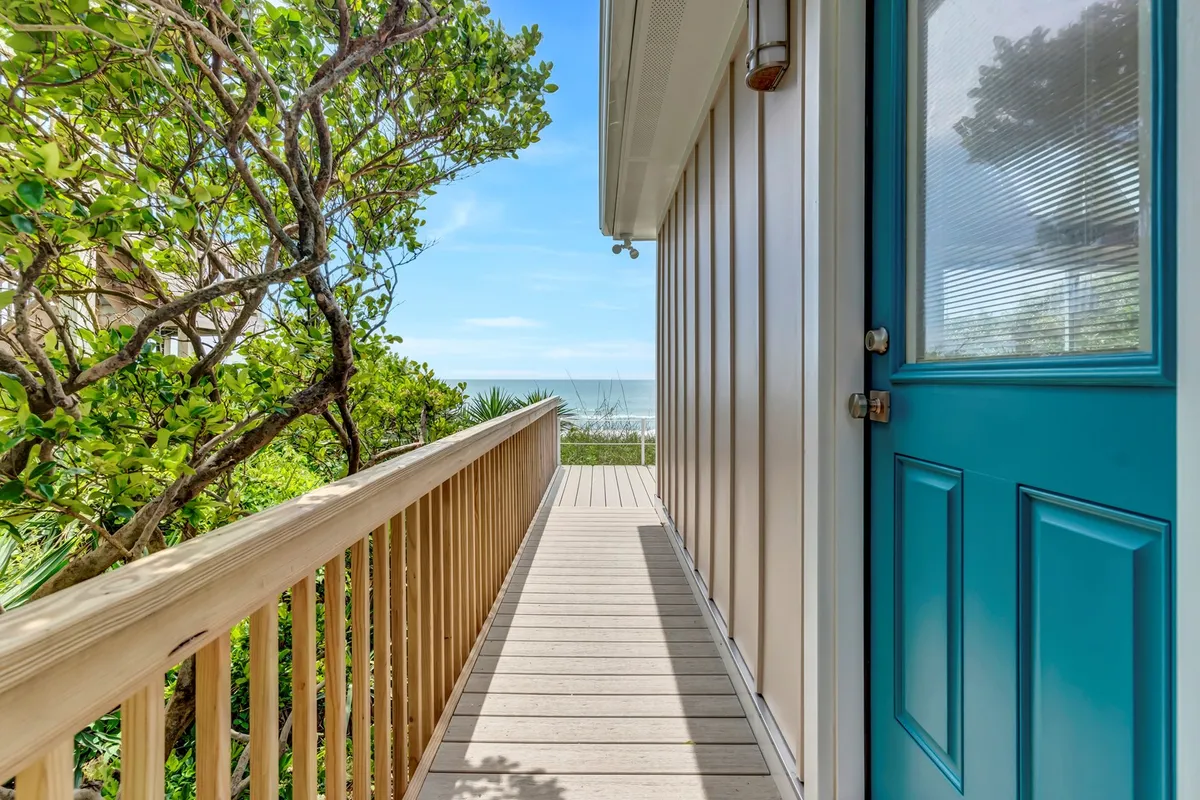 This exterior shot shows a blue door opening to a wooden walkway framed by lush greenery, with an inviting ocean view just beyond. Someone could easily be imagined walking a dog towards the beach or relaxing on the deck, soaking in the coastal atmosphere.