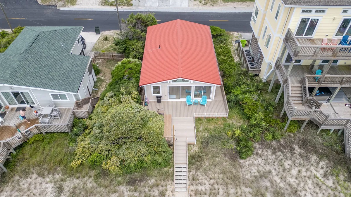 A charming red-roofed beach cottage with a wooden deck furnished with blue chairs, facing the beach. The setting hints at family gatherings, morning coffees, and evening relaxation with sweeping ocean views.