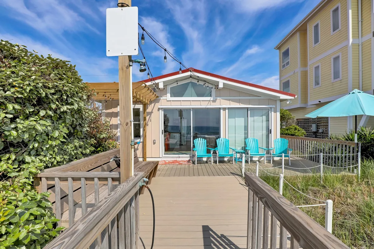 A cheerful beachfront patio with blue Adirondack chairs facing the ocean, a wooden walkway, and a sunny, open space that invites relaxation and social gatherings. The scene evokes a sense of comfort and casual enjoyment right by the water.