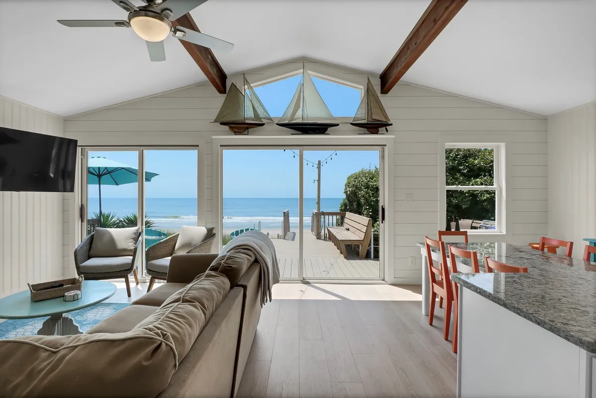 A bright, open-plan living and dining area with large windows framing ocean views. The space includes a light-colored sofa, a sizable dining table, and coastal sailing decor, all bathed in natural light, creating a tranquil and inviting atmosphere for gatherings.