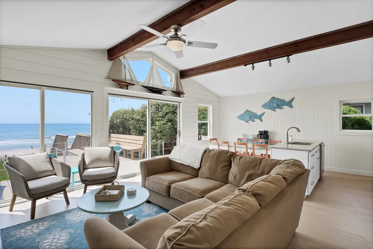 A welcoming open-plan living and dining area, with a comfortable sofa, ocean views through large windows, dining space, and playful fish-themed artwork. The ceiling fan and light-filled room enhance the breezy, relaxed atmosphere, ideal for socializing or unwinding.