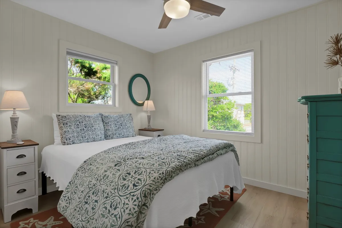 A serene queen bedroom with patterned bedding, a nightstand with a warm lamp, and a green mirror. Sunlight streams in from windows overlooking lush greenery, creating a tranquil, restful atmosphere.