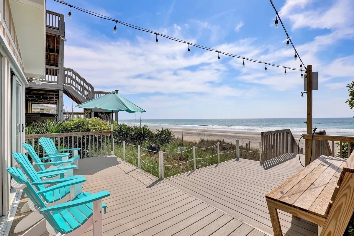 A beachfront deck is adorned with turquoise chairs and string lights, overlooking a stunning ocean vista. The area is set up for outdoor dining or lounging, encouraging guests to enjoy meals, drinks, or conversations against a backdrop of sea and sky. The presence of string lights hints at lively evenings spent together.