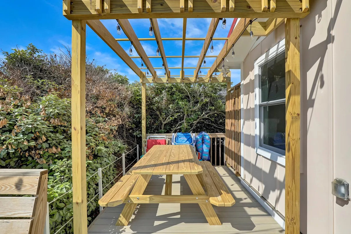 A cozy outdoor dining area beneath a wooden pergola, featuring a picnic table and hanging lights, surrounded by greenery. The space exudes a welcoming, communal vibe ideal for meals and gatherings.