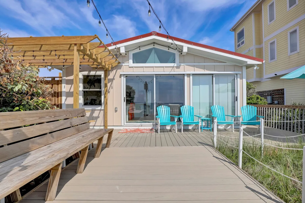 A spacious oceanfront deck with blue chairs, a pergola, and glass doors leading inside. The setup is perfect for lounging, outdoor dining, and enjoying expansive views of the water.