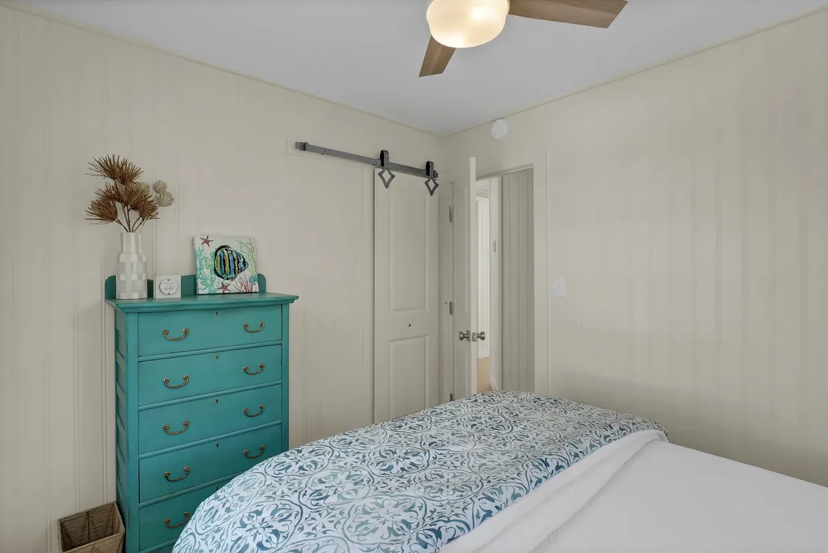 A cozy bedroom with a turquoise dresser, floral patterned bedding, and a ceiling fan. The doorway offers a glimpse into another room, suggesting the cottage’s spaciousness and flow. The setup feels restful and inviting, perfect for a peaceful night or quiet reading time.