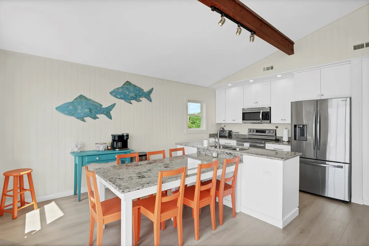 A playful, family-friendly kitchen and dining space with a granite countertop, colorful chairs, and blue fish artwork. The setup invites gatherings for meals, casual breakfasts, or entertaining, while maintaining a vibrant coastal vibe.