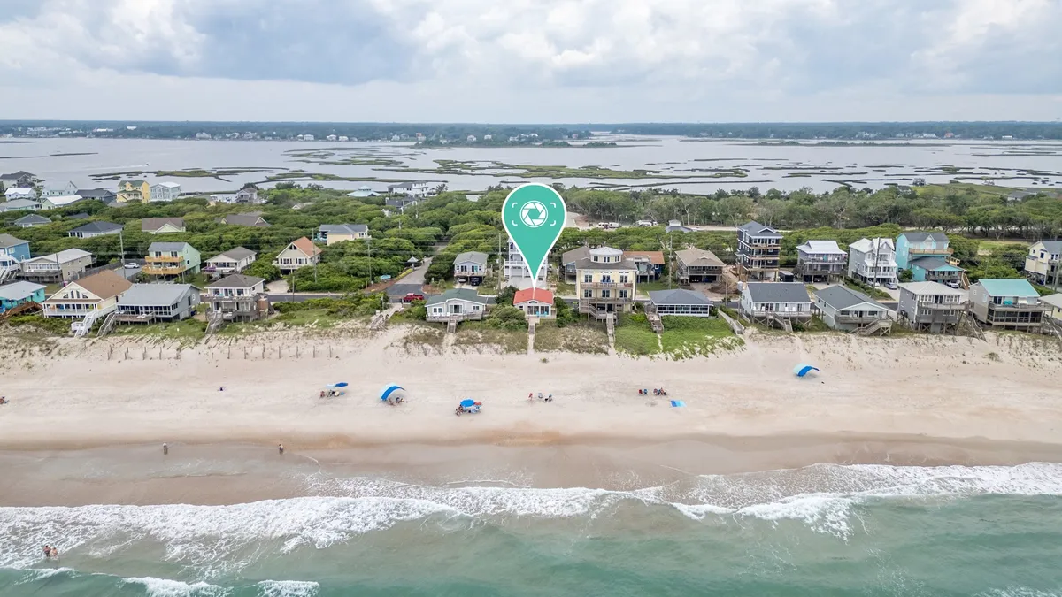 This image features a beachfront property with the ocean waves in the background, colorful beach umbrellas dotting the shore, and surrounding houses. The scene includes people sunbathing, swimming, walking along the sand, and enjoying family gatherings, all set against a relaxed coastal backdrop.