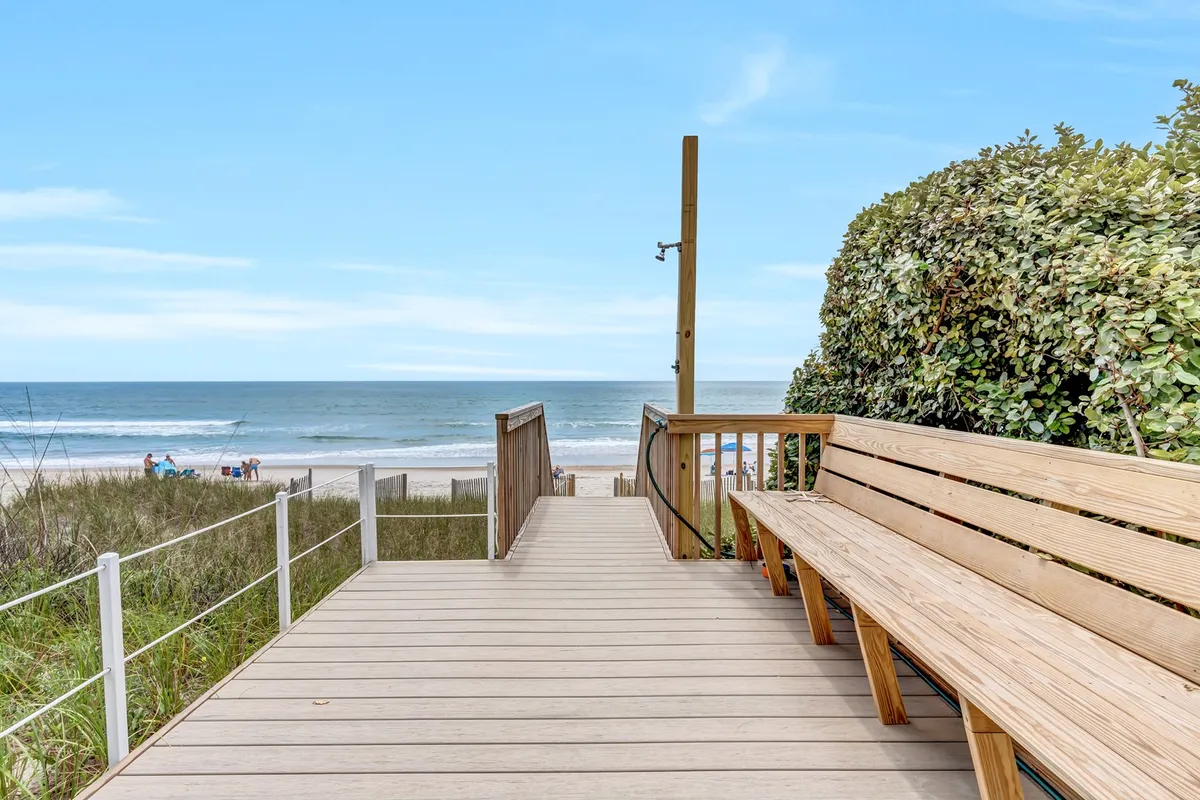 A serene scene showing a wooden walkway leading to the beach, with a seating area overlooking the ocean. The view highlights easy private access to the sand and inviting spaces to enjoy the coastal scenery.