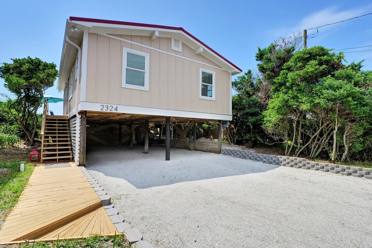 An elevated coastal cottage with wide wooden steps and lush greenery around its base. The house exudes a welcoming aura, and the steps and porch provide an inviting space for morning coffee or evening gatherings. The setting is private yet open, perfect for both relaxation and social interaction.