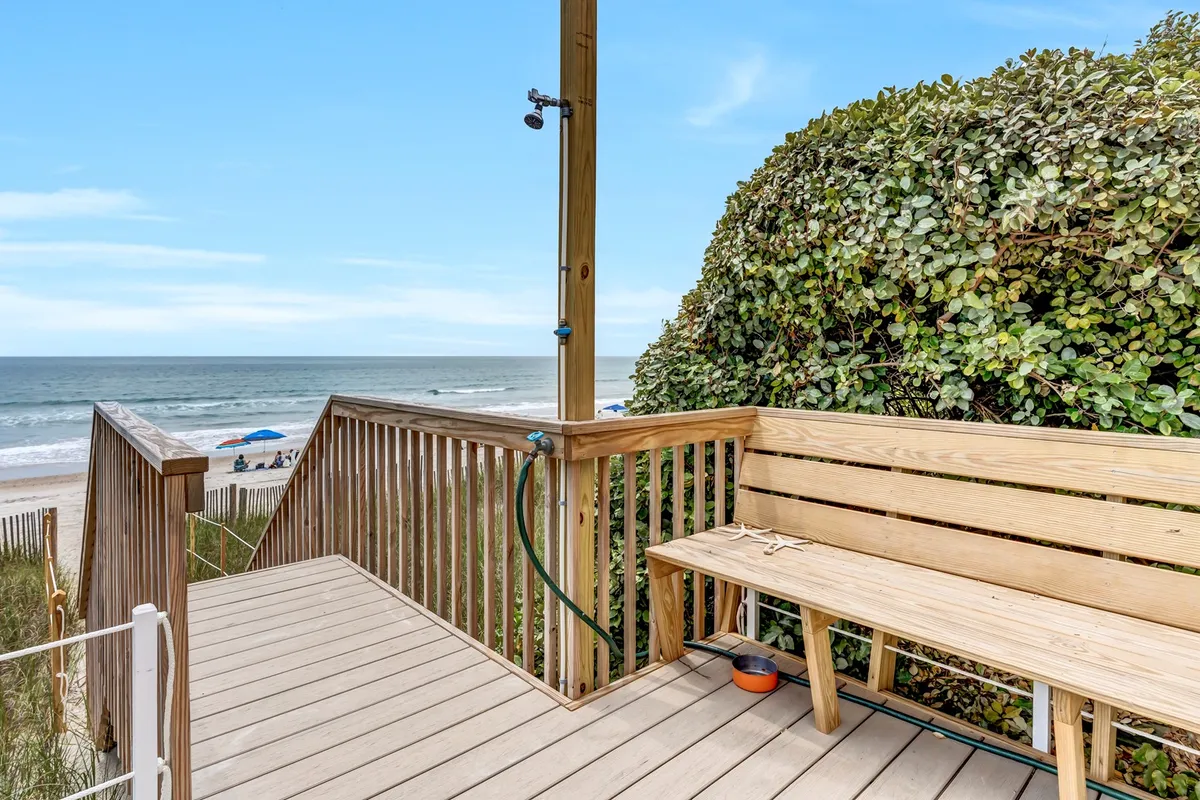 A wooden bench sits amidst lush green foliage, facing an expansive ocean view. A sandy path leads from the garden area toward the beach, evoking a sense of tranquility and easy access to the shoreline. The scene feels welcoming for groups to gather and enjoy the seaside breeze, perfect for relaxing, chatting, or watching the waves together.